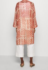 Lång, lös kimono i rosa tyg med guldaccent och geometriska mönster. Har trekvartsärmar och sidofickor, kombineras med vita, korta byxor.