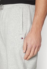 Pantalones deportivos de algodón gris con bolsillos laterales y un pequeño logo bordado en rojo, blanco y azul cerca de la cintura. Se aprecia una textura suave.