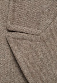 Gros plan sur le revers d'un manteau en laine marron montrant un tissu texturé et un col cranté soigneusement plié.