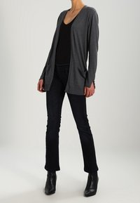 Grå, långärmad kofta i mjukt tyg med draperad framkant, två framfickor och en avslappnad passform, kombinerad med mörka jeans och ankelboots.