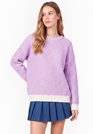 Woman wearing a light purple knit sweater with white cuffs and hem paired with a blue pleated skirt, standing with one hand on hip.