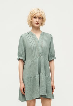 Γυναίκα με σγουρά ξανθά μαλλιά, που φοράει ένα χαλαρό, υφασμάτινο φόρεμα σε φουντουκί πράσινο με τρία κουμπιά και πιέτες μπροστά.