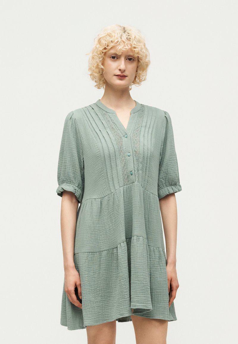 Γυναίκα με σγουρά ξανθά μαλλιά, που φοράει ένα χαλαρό, υφασμάτινο φόρεμα σε φουντουκί πράσινο με τρία κουμπιά και πιέτες μπροστά.