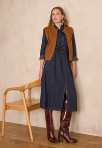 Robe en denim avec une taille cintrée, une fente à l'avant et un gilet en tricot marron. Bottes à talons bordeaux hautes. Chaise en bois en arrière-plan sur un sol en bois.