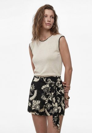 Kvinde med bølget hår iført ærmeløs beige top med sort kant og sort blomster wrap-nederdel, stående med hænderne foldet bag ryggen.