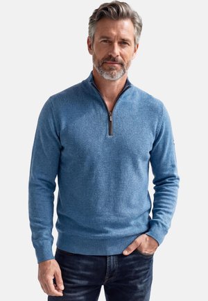 Mittelalter Mann mit grauem Haar und Bart, der einen blauen, strukturierten Pullover mit Viertel-Reißverschluss und dunkle Jeans trägt, steht mit einer Hand in der Tasche.