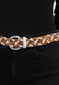 Bruin-witte geweven leren riem met zilverkleurige ronde gesp en een rechthoekige gespbeschermer, gedragen over zwarte kleding.