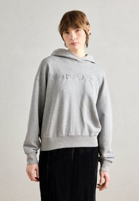 Grå hettegenser laget av mykt stoff, med en avslappet passform og preget "Marc O'Polo"-tekst på fronten. Båret med et svart plisserte skjørt.