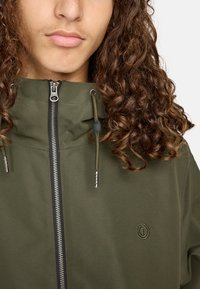 Olijfgroen jack met hoge kraag, rits aan de voorkant en trekkoordkap. Bevat zilverkleurige details en een getextureerd logo op de borst.