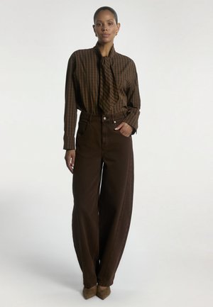 Vrouw draagt een bruin gestreepte blouse met een nekstrik, hoog getailleerde, wijd uitlopende donkerbruine broek, bruine puntige schoenen, staand tegen een witte achtergrond.
