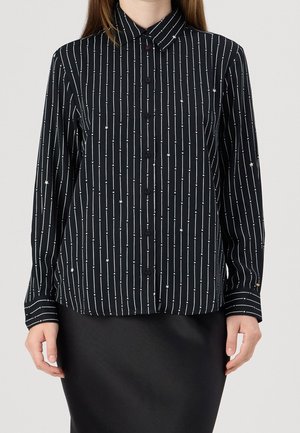 Zwarte blouse met knopen, witte verticale strepen en kleine sterpatronen, gedragen met een zwarte rok, getoond van nek tot heupen.