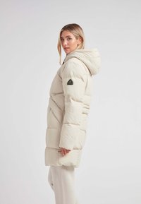 Chaqueta acolchada en blanco roto, con capucha, costuras diagonales y un parche con el logo en la manga. Material suave e insulated, diseño hasta la rodilla.