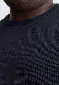 Dunkelblauer, gestrickter Pullover mit einem gerippten Rundhalsausschnitt und subtilen Logodetails in weicher Textur. Nahaufnahme des Kleidungsstücks.