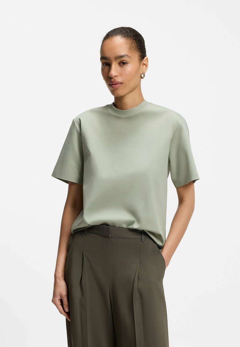 Femme les cheveux attachés portant une chemise vert clair à manches courtes et un pantalon plissé taille haute vert foncé, debout avec une main dans la poche.