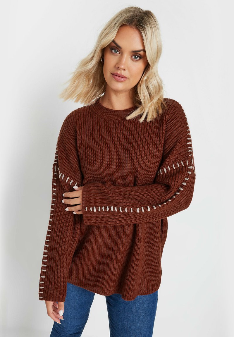 Brun stickad tröja med långa ärmar, med vit dekorativ sömnad längs ärmarna. Buren med blå jeans, som visar en lös passform.