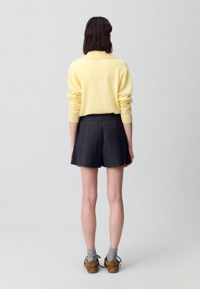 Suéter de color amarillo claro con cuello acanalado, combinado con pantalones cortos oscuros. El modelo lleva calcetas grises y zapatos marrones. Fondo liso.