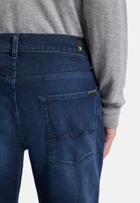 Jeans in denim blu con una tasca posteriore caratterizzata da un delicato dettaglio di cucitura a onda. La cintura è scura e presenta una piccola etichetta nera.
