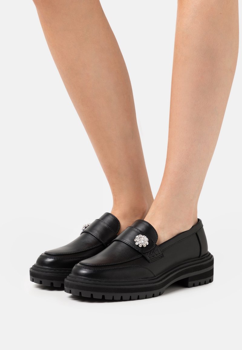 Mocassins noirs épais avec semelles épaisses et broches décoratives en strass, portés pieds nus sur fond blanc.
