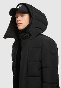 Chaqueta acolchada negra con una capucha estructurada, que presenta una textura suave y cierres de presión. Un parche de logo negro se encuentra en el hombro izquierdo.