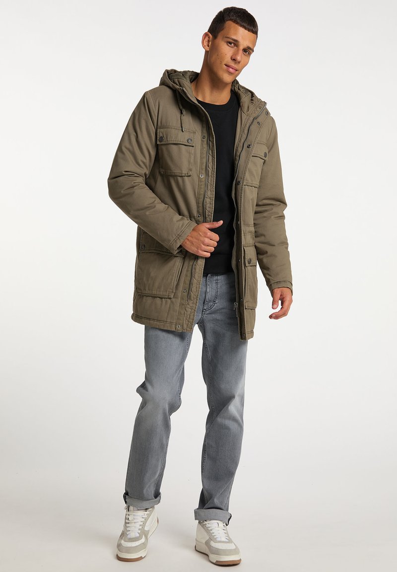 Parka giacca verde oliva con cappuccio, tasche frontali e dettagli con bottoni. Indossata sopra una maglietta nera e abbinata a jeans azzurri chiari e sneakers.