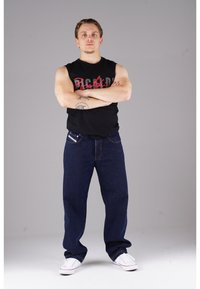 Schwarzes ärmelloses Shirt, blaue Jeans, weiße Schuhe. Die Jeans haben einen geraden Schnitt und sichtbare Nähte entlang der Taschen.