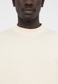 Világos bézs crew-neck póló, puha pamutból készült. Simaszerkezetű, minimalista dizájnnal és díszítés nélküli nyakkal rendelkezik.