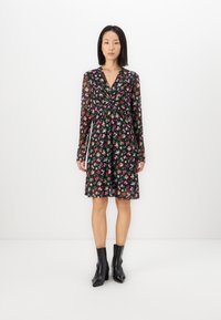 Robe noire à fleurs avec des manches longues, dotée d'un détail en devant noué, d'un motif floral multicolore et d'un ourlet évasé, assortie à des bottes noires à la cheville.