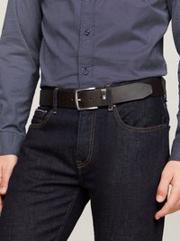 Hombre que lleva un pantalón vaquero azul oscuro con costuras naranjas, un cinturón de cuero negro con hebilla plateada y una camisa de puntos azul marino.
