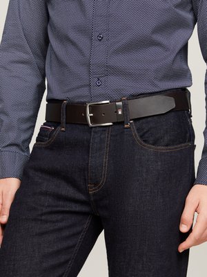 Un homme portant un jean bleu foncé avec des coutures orange, une ceinture en cuir noire avec une boucle en argent et une chemise bleu marine à pois.