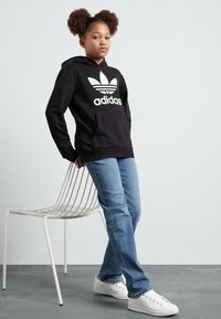 Musta kapuutsiga särk, millel on valge Adidas'i logo ja kolm triipu, kombineerituna siniste teksade ja valgete tossudega, helesinise halli tausta ees.