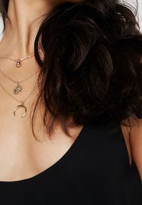 Les colliers en or plaqué comportent un pendentif rond, un charme circulaire texturé et une pièce en forme de croissant, portés sur un haut noir.