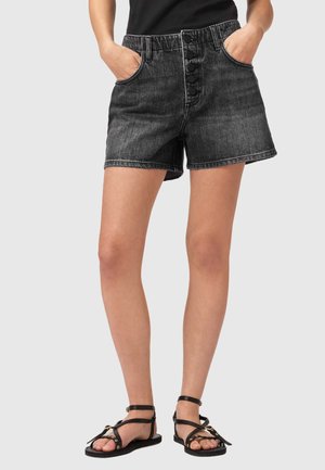 Kvinde iført sorte denimshorts med høj talje og knaplukning, hænderne i lommerne, kombineret med sorte flade sandaler med stropper mod en grå baggrund.