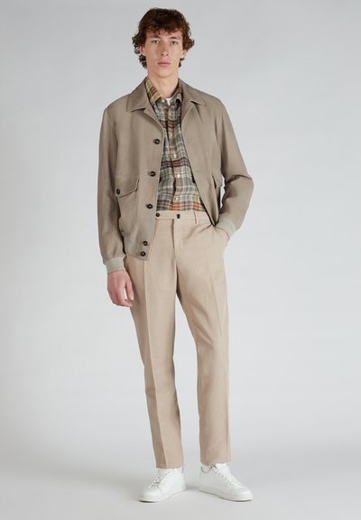 Giacca leggera beige con chiusura a bottoni e due tasche, indossata sopra una camicia a quadri multicolore, abbinata a pantaloni beige chiaro e sneakers bianche.
