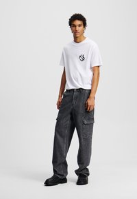 Hvid T-shirt med sort logo, parret med oversized mørkegrå cargo bukser med sidelommer, tekstureret stof og sorte fodtøj.