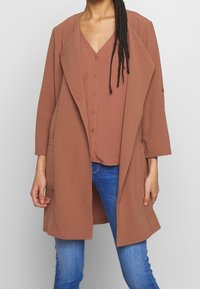 Rostfärgad lång kappa med sjalkrage, sidfickor och öppen framsida, över en matchande topp, tillsammans med blå jeans.
