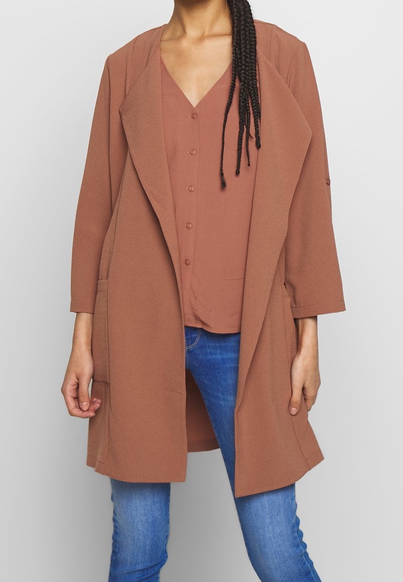 Rostfärgad lång kappa med sjalkrage, sidfickor och öppen framsida, över en matchande topp, tillsammans med blå jeans.