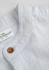 Jasnoniebieski, żebrowany kołnierzyk koszuli z brązowym guzikiem. Etykieta informuje "CODRILLO", rozmiar 140, z teksturowanym detalem powierzchni.