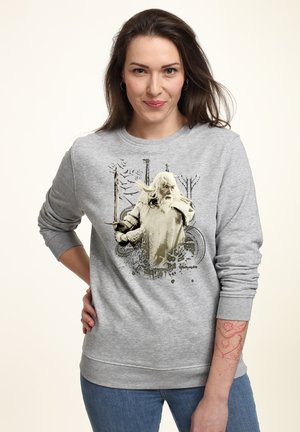 Kvinde iført grå sweatshirt med grafik af en ældre mand, der svinger et sværd, samt abstrakte elementer af natur og våben.