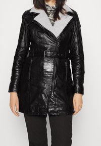 Chaqueta de cuero negra con cremallera frontal, amplio cuello y suave forro gris. Presenta una cintura con cinturón y dos bolsillos laterales con cremallera.