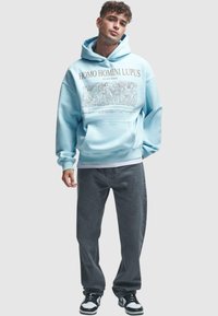 Light blue hoodie with a graphic print featuring "HOMO HOMINI LUPUS" and figures. Paired with gray pants and black and white sneakers.