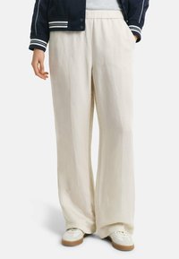 Helle beige weit geschnittene Hose aus einem strukturierten Stoff, mit einem elastischen Taillenbund und lockerer Passform. Kombiniert mit weißen Sneakers.