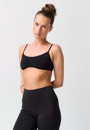 Woman with hair in a bun wearing a black sports bra and high-waisted black leggings, standing against a plain light background.