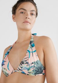 Mulher a usar um biquíni halter rosa com padrão de folha e flor tropical em azul e verde, contra um fundo branco liso.