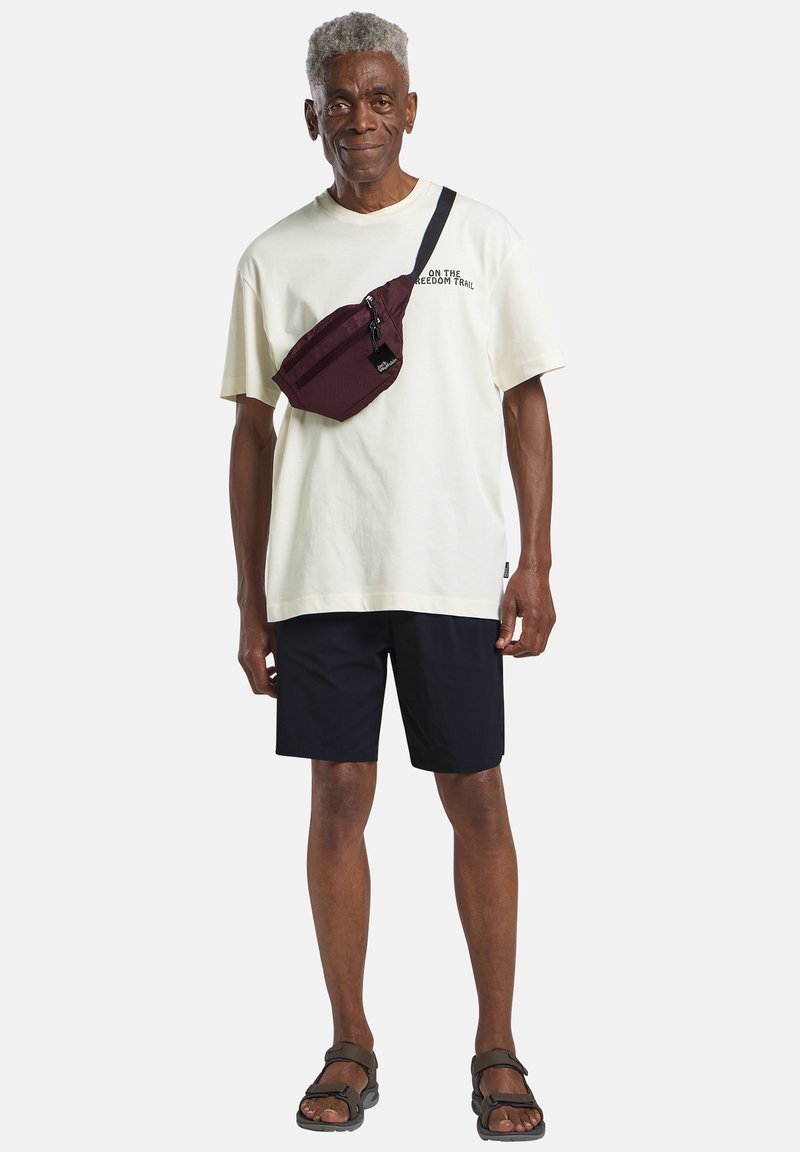 Borsa a tracolla bordeaux, t-shirt di cotone color crema, pantaloni corti scuri, indossando sandali neri. La borsa presenta una cerniera e una tracolla regolabile.