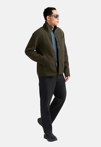 Olivgrüne Rippenjacke mit Stehkragen und Reißverschluss, kombiniert mit einem blauen, geschichteten Top und schwarzen Hosen. Das Modell trägt eine Sonnenbrille und schwarze Schuhe.