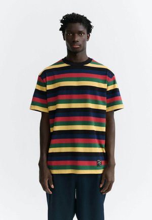 Homme portant un T-shirt à manches courtes avec des rayures horizontales jaune, rouge, verte, bleu marine et noire, se tenant devant un fond blanc.