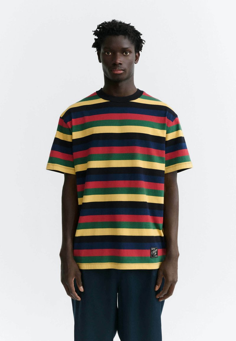 Homme portant un T-shirt à manches courtes avec des rayures horizontales jaune, rouge, verte, bleu marine et noire, se tenant devant un fond blanc.