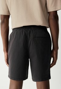 Pantaloni shorts neri in cotone con vita elasticizzata, dotati di una tasca posteriore e orlo arrotolato. Il tessuto ha una texture opaca.