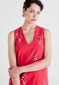 Red sleeveless dress with a V-neck, embellished with floral sequins in pink, purple, and green, featuring a smooth, lightweight texture.