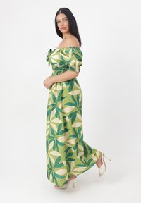 Grön blommig maxiklänning med off-shoulder halsringning, smockad midja och stort bladmönster. Materialet är lätt och har en flödande silhuett.